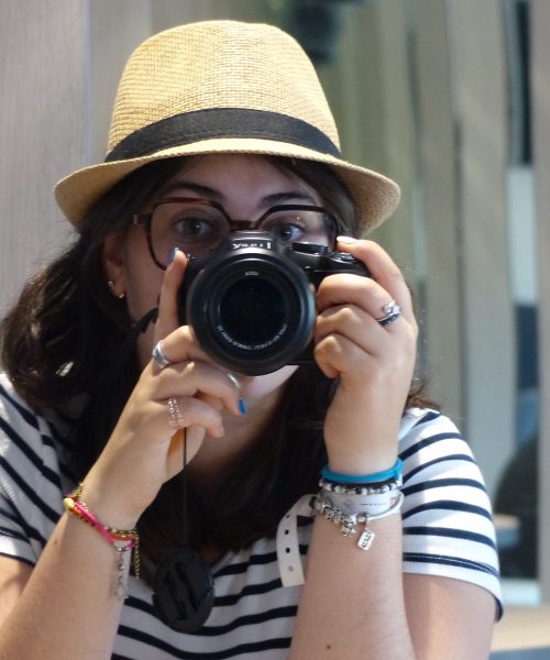 Agathe en train de se prendre en photo dans un miroir avec un appareil photo. Elle porte un chapeau de paille et un tee shirt marinière